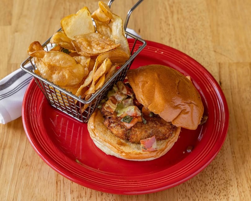 A chicken sandwich with salsa and a side of potato chips in a metal basket on a red plate.