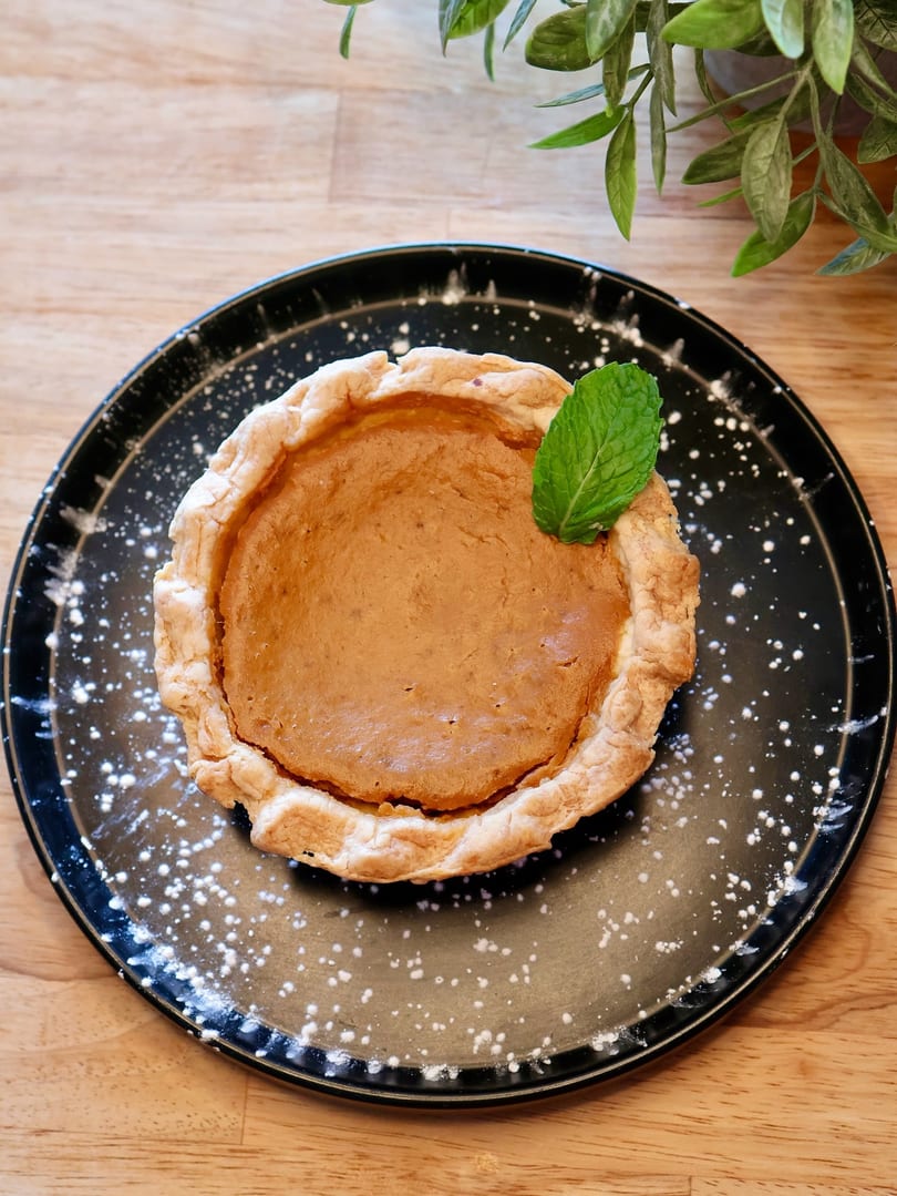 A mini pumpkin pie with a mint leaf on a black plate dusted with powdered sugar.