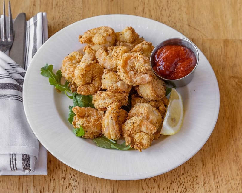 Crispy fried shrimp on a white plate with arugula, cocktail sauce, and a lemon wedge.