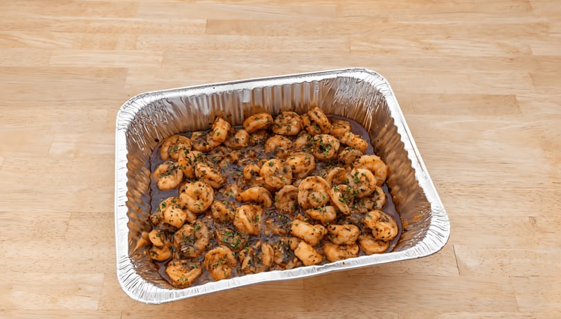 Shrimp scampi in a foil pan, garnished with parsley, on a light wooden table.